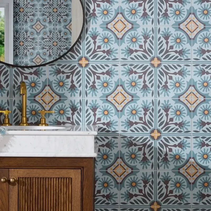 Close-up of Indirah Porcelain Azure tiles showing a repeating geometric pattern with floral motifs in muted blue, grey and warm accent tones.
