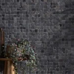 Enso Cobble Cube Charcoal wall dark grey mosaic tiles with tonal shifts and an organic, uneven surface, creating a textured backdrop paired with warm metal and soft greenery accents.