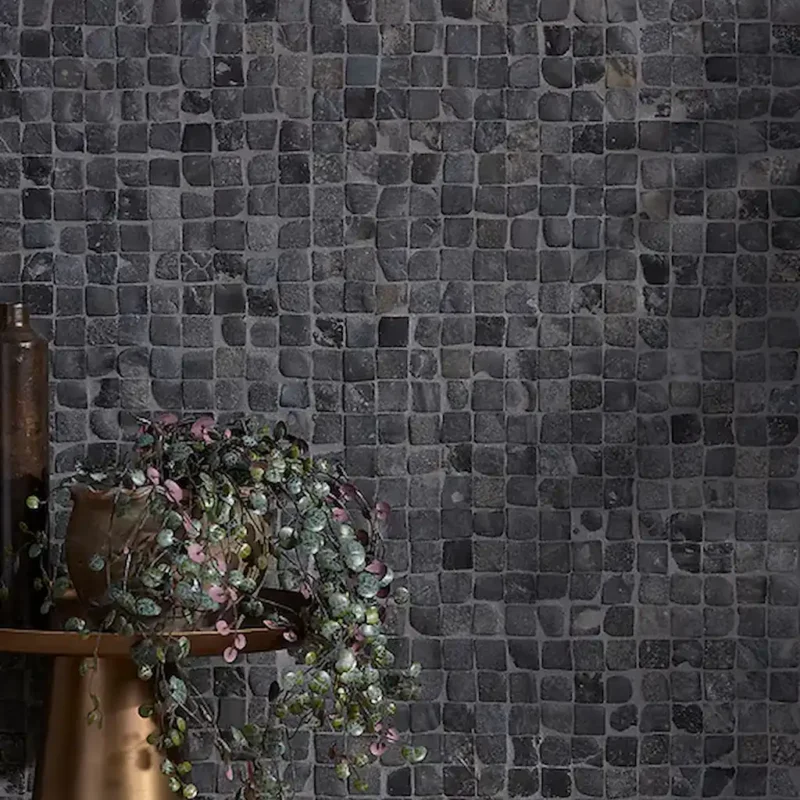 Enso Cobble Cube Charcoal wall dark grey mosaic tiles with tonal shifts and an organic, uneven surface, creating a textured backdrop paired with warm metal and soft greenery accents.